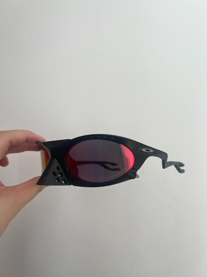 Może przedstawiać: Czarne okulary przeciwsłoneczne z czerwonymi soczewkami. Okulary mają unikalny, kanciasty design z zakrzywionym zausznikiem. Soczewki są w kolorze głębokiej czerwieni, a oprawki czarne.