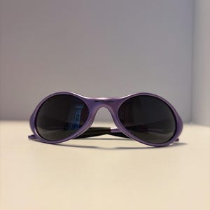 K&ouml;nnte beinhalten: Violette Sonnenbrille mit dunklen Gl&auml;sern. Die Sonnenbrille hat einen abgerundeten Rahmen und einen Steg. Die B&uuml;gel sind d&uuml;nn und verbinden sich mit dem Rahmen. Die Sonnenbrille liegt auf einer wei&szlig;en Oberfl&auml;che.