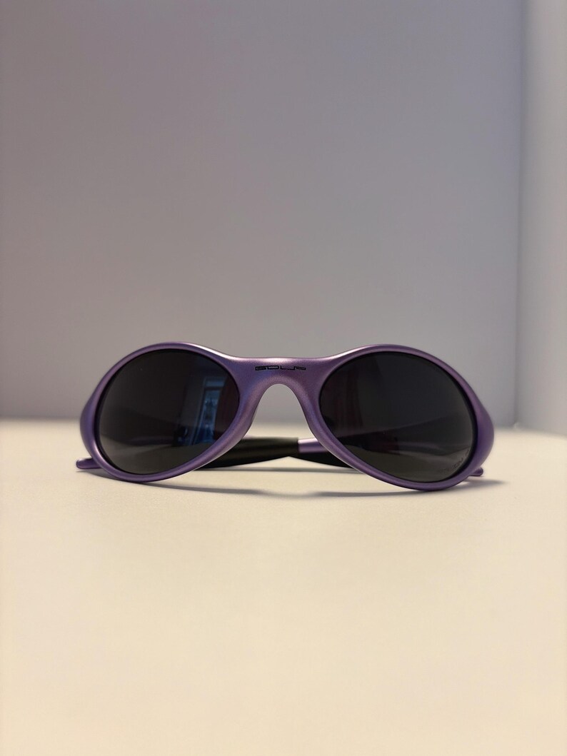 K&ouml;nnte beinhalten: Violette Sonnenbrille mit dunklen Gl&auml;sern. Die Sonnenbrille hat einen abgerundeten Rahmen und einen Steg. Die B&uuml;gel sind d&uuml;nn und verbinden sich mit dem Rahmen. Die Sonnenbrille liegt auf einer wei&szlig;en Oberfl&auml;che.