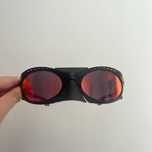 Może przedstawiać: Czarne sportowe okulary przeciwsłoneczne z czerwonymi soczewkami. Okulary mają owinięty wok&oacute;ł design z mostkiem i g&oacute;rnym paskiem łączącym dwie soczewki. Soczewki są w żywym czerwonym kolorze, a oprawki czarne.
