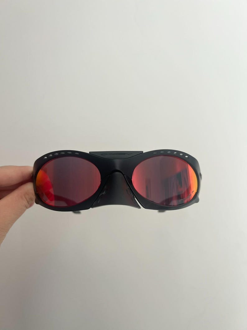 Może przedstawiać: Czarne sportowe okulary przeciwsłoneczne z czerwonymi soczewkami. Okulary mają owinięty wok&oacute;ł design z mostkiem i g&oacute;rnym paskiem łączącym dwie soczewki. Soczewki są w żywym czerwonym kolorze, a oprawki czarne.