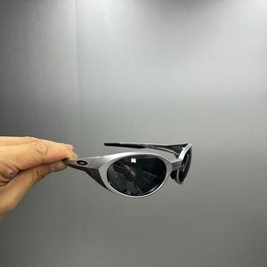 Pode incluir: &Oacute;culos de sol com arma&ccedil;&atilde;o prateada e lentes escuras. Os &oacute;culos de sol t&ecirc;m um design elegante e moderno com uma arma&ccedil;&atilde;o curva e uma est&eacute;tica desportiva. As lentes s&atilde;o escuras, proporcionando sombra e prote&ccedil;&atilde;o solar.