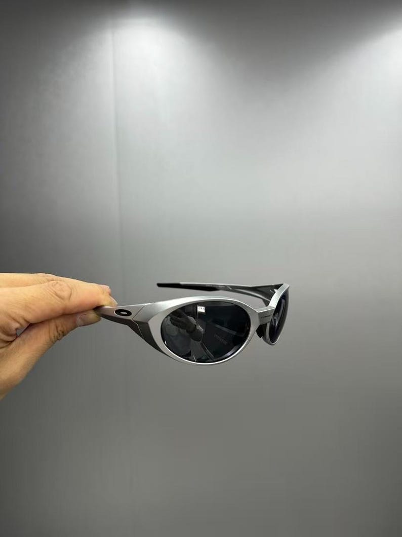 Pode incluir: &Oacute;culos de sol com arma&ccedil;&atilde;o prateada e lentes escuras. Os &oacute;culos de sol t&ecirc;m um design elegante e moderno com uma arma&ccedil;&atilde;o curva e uma est&eacute;tica desportiva. As lentes s&atilde;o escuras, proporcionando sombra e prote&ccedil;&atilde;o solar.