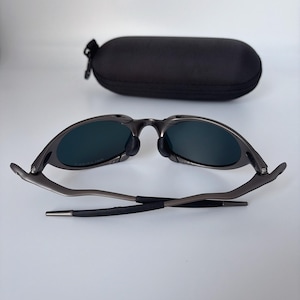 May include: Dark grey sunglasses with dark lenses and a black case. The sunglasses are open, showing the lenses and frame. The case is oval and has a zip.