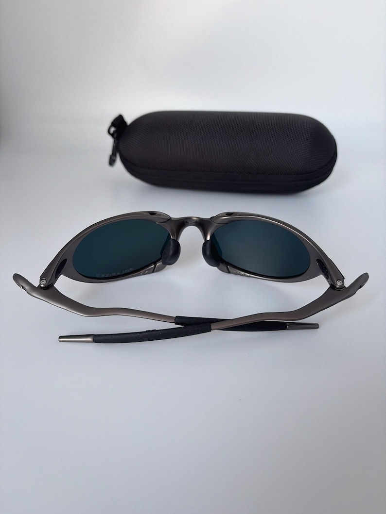 May include: Dark grey sunglasses with dark lenses and a black case. The sunglasses are open, showing the lenses and frame. The case is oval and has a zip.