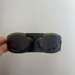 May include: Olive green sunglasses with dark lenses. The sunglasses have a unique design with a bridge connecting the two lenses. The frames have small, decorative details. The sunglasses are held against a white background.