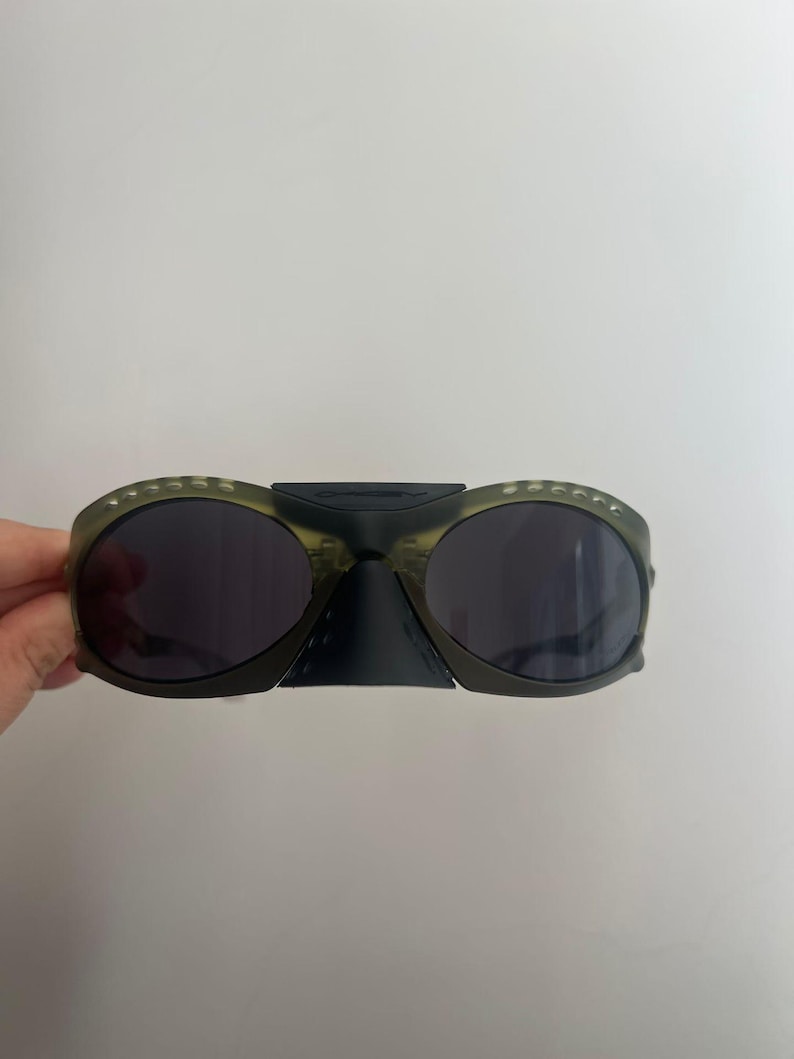 May include: Olive green sunglasses with dark lenses. The sunglasses have a unique design with a bridge connecting the two lenses. The frames have small, decorative details. The sunglasses are held against a white background.