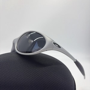 Puede incluir: Gafas de sol plateadas con lentes oscuras sobre un estuche negro texturizado. Las gafas tienen un dise&ntilde;o moderno con patillas curvas y un peque&ntilde;o logotipo. El estuche est&aacute; hecho de un material tejido duradero.