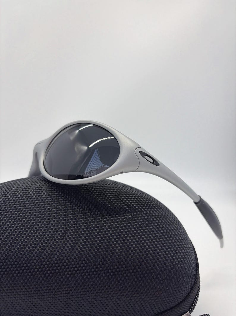 Puede incluir: Gafas de sol plateadas con lentes oscuras sobre un estuche negro texturizado. Las gafas tienen un dise&ntilde;o moderno con patillas curvas y un peque&ntilde;o logotipo. El estuche est&aacute; hecho de un material tejido duradero.
