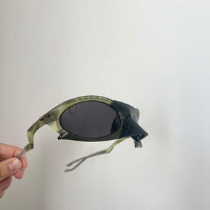 May include: Olive green and black sunglasses with dark lenses. The sunglasses have a unique, angular design with a textured nose bridge and temples. The frame has a camouflage-like pattern. The arms are a translucent green.