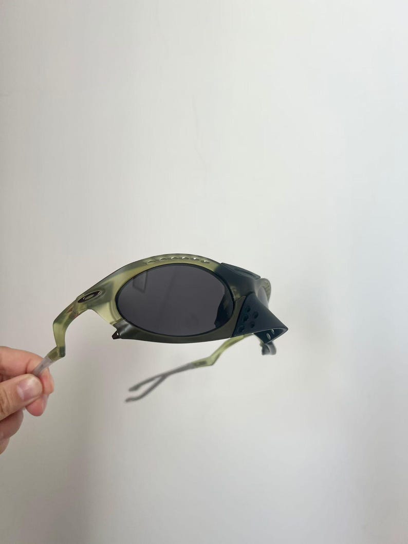 May include: Olive green and black sunglasses with dark lenses. The sunglasses have a unique, angular design with a textured nose bridge and temples. The frame has a camouflage-like pattern. The arms are a translucent green.