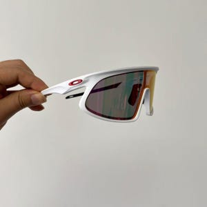 May include: White sports sunglasses with a red logo on the frame. The lenses have a gradient of colors, including red, orange, and purple. The sunglasses are held by a hand against a plain background.