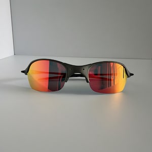 May include: Black sunglasses with a rimless design and vibrant red-orange mirrored lenses. The sunglasses have a sleek, modern aesthetic, suitable for outdoor activities. The lenses reflect a gradient of colors.
