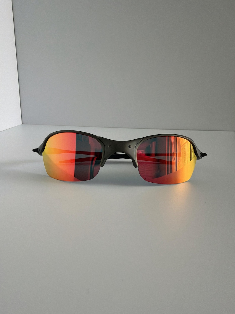 May include: Black sunglasses with a rimless design and vibrant red-orange mirrored lenses. The sunglasses have a sleek, modern aesthetic, suitable for outdoor activities. The lenses reflect a gradient of colors.