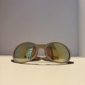 May include: A pair of beige sunglasses with oval lenses and a thick frame. The lenses have a reflective, golden-yellow tint. The sunglasses are resting on a white surface.