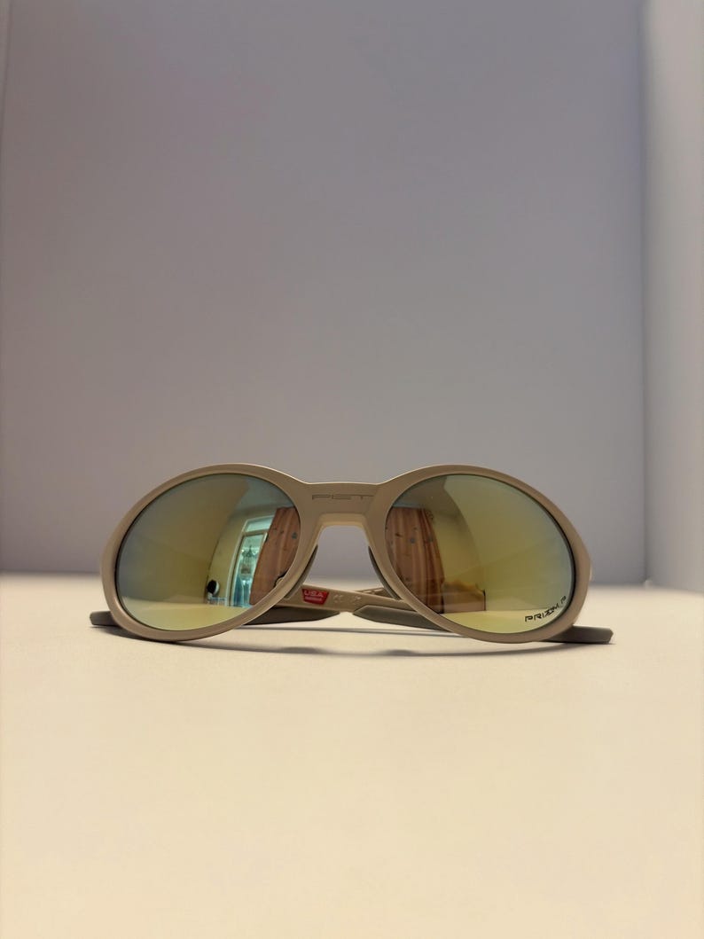 May include: A pair of beige sunglasses with oval lenses and a thick frame. The lenses have a reflective, golden-yellow tint. The sunglasses are resting on a white surface.
