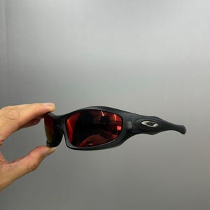 May include: Black sunglasses with red lenses. The sunglasses have a sleek, curved design with a matte finish. The temples feature a small, silver-coloured emblem. The lenses reflect the surrounding environment.