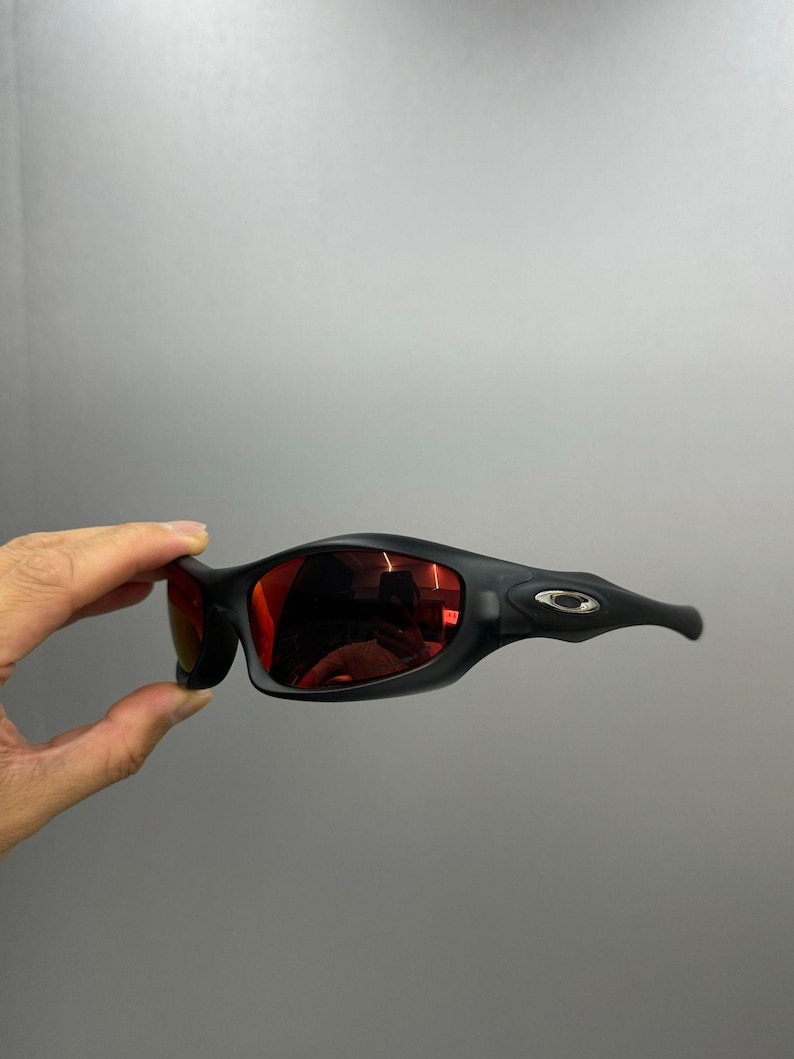 May include: Black sunglasses with red lenses. The sunglasses have a sleek, curved design with a matte finish. The temples feature a small, silver-coloured emblem. The lenses reflect the surrounding environment.