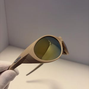 May include: Tan-colored sunglasses with a curved frame and reflective lenses. The sunglasses have a sleek, modern design. The text "FIELD GEAR" is printed on the arm of the sunglasses.