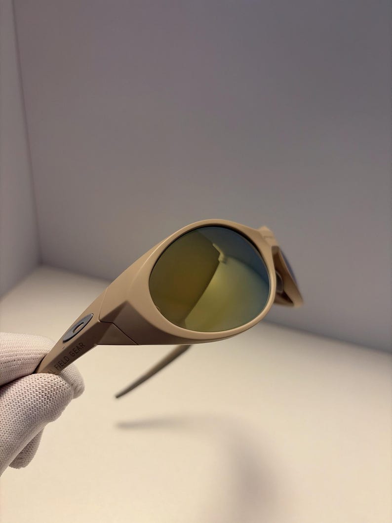 May include: Tan-colored sunglasses with a curved frame and reflective lenses. The sunglasses have a sleek, modern design. The text "FIELD GEAR" is printed on the arm of the sunglasses.