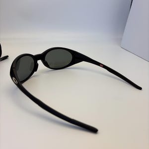 May include: Black oval sunglasses with dark gray lenses. The sunglasses have a sleek, modern design with a thin frame and curved temples. A small red and white USA logo is visible on the temple.