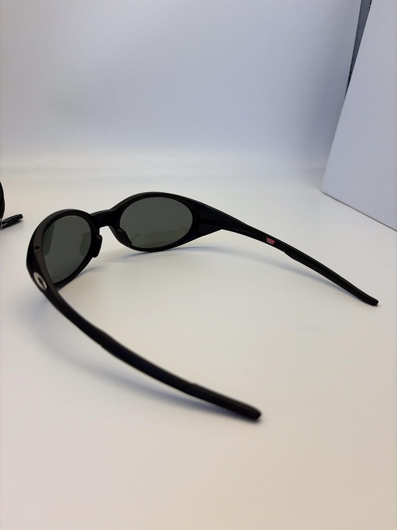 May include: Black oval sunglasses with dark gray lenses. The sunglasses have a sleek, modern design with a thin frame and curved temples. A small red and white USA logo is visible on the temple.