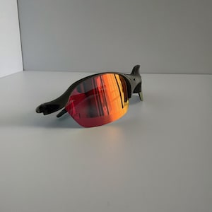 May include: Black sunglasses with red and orange mirrored lenses. The sunglasses have a sleek, modern design with a curved frame and a streamlined shape. The lenses reflect a gradient of colors.
