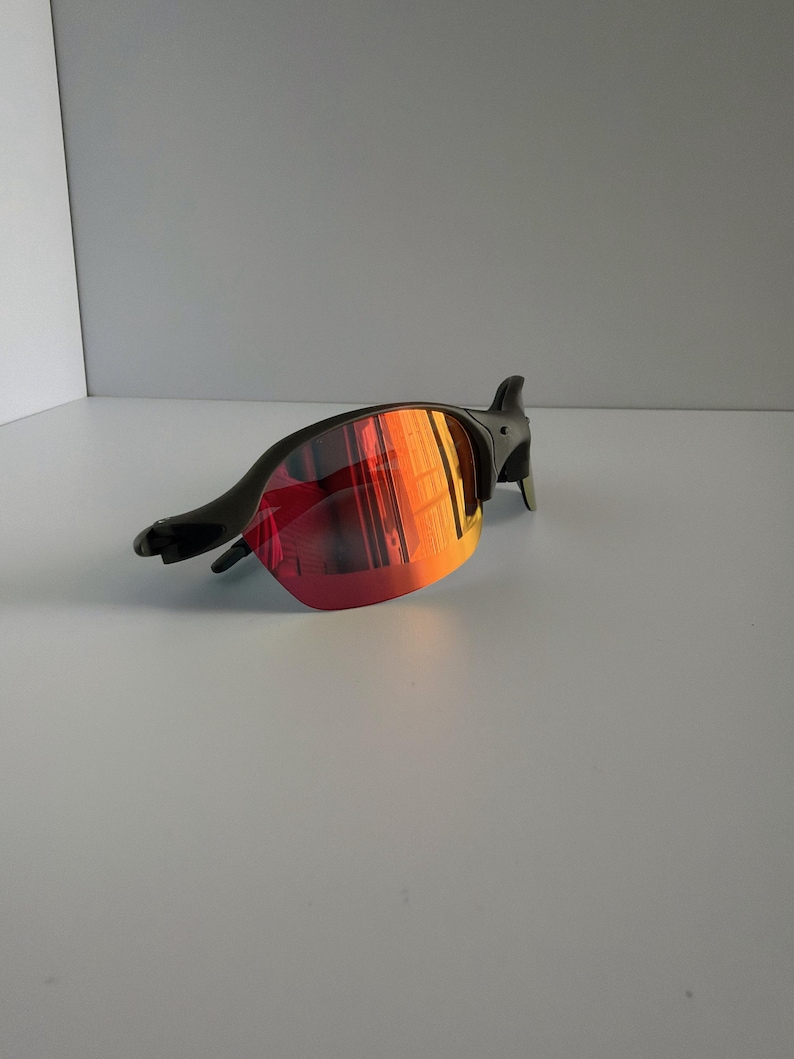 May include: Black sunglasses with red and orange mirrored lenses. The sunglasses have a sleek, modern design with a curved frame and a streamlined shape. The lenses reflect a gradient of colors.