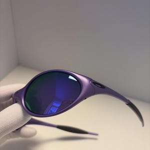 May include: Purple sunglasses with blue mirrored lenses. The sunglasses have a rounded frame and black temples. The lenses reflect a blue hue. The sunglasses are held by a gloved hand.