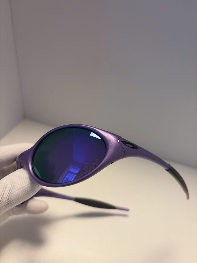 May include: Purple sunglasses with blue mirrored lenses. The sunglasses have a rounded frame and black temples. The lenses reflect a blue hue. The sunglasses are held by a gloved hand.