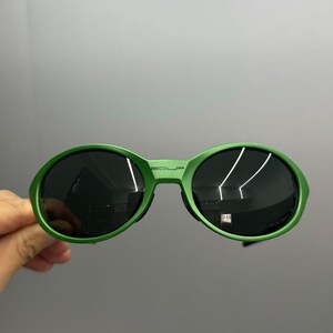Może przedstawiać: Zielone okulary przeciwsłoneczne z okrągłymi soczewkami i pasującą oprawką. Okulary przeciwsłoneczne mają ciemne soczewki, a na g&oacute;rze oprawki wydrukowano napis "SOLP". Okulary są trzymane na szarym tle.