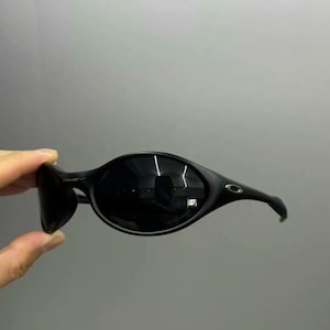 Puede incluir: Gafas de sol negras con montura curvada de forma ovalada. Las lentes son oscuras y las patillas se curvan alrededor de las orejas. Las gafas de sol est&aacute;n sujetas por una mano sobre un fondo gris.