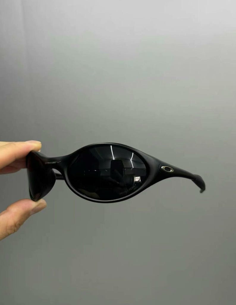 Puede incluir: Gafas de sol negras con montura curvada de forma ovalada. Las lentes son oscuras y las patillas se curvan alrededor de las orejas. Las gafas de sol est&aacute;n sujetas por una mano sobre un fondo gris.