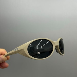 May include: Tan-colored sunglasses with oval-shaped lenses and a curved frame. The lenses are dark, and the arms of the sunglasses are thin. The sunglasses are held by a hand against a gray background.