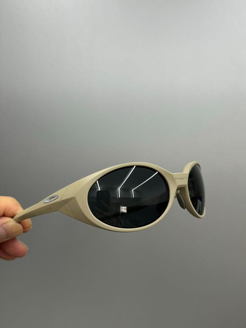 May include: Tan-colored sunglasses with oval-shaped lenses and a curved frame. The lenses are dark, and the arms of the sunglasses are thin. The sunglasses are held by a hand against a gray background.