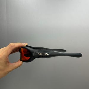 May include: Black sports sunglasses with red lenses. The sunglasses have a sleek design with a matte finish. The frame features a small, silver logo on the temples. The lenses are a vibrant red colour.
