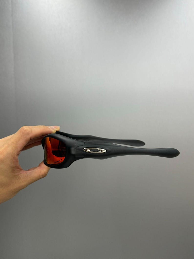 May include: Black sports sunglasses with red lenses. The sunglasses have a sleek design with a matte finish. The frame features a small, silver logo on the temples. The lenses are a vibrant red colour.