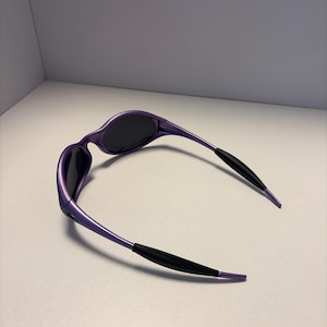 K&ouml;nnte beinhalten: Violette Sonnenbrille mit dunklen Gl&auml;sern und schwarzen B&uuml;geln. Die Sonnenbrille hat ein schlankes, geschwungenes Design. Die Fassungen sind in einem leuchtenden Violett gehalten, vor einem neutralen Hintergrund.