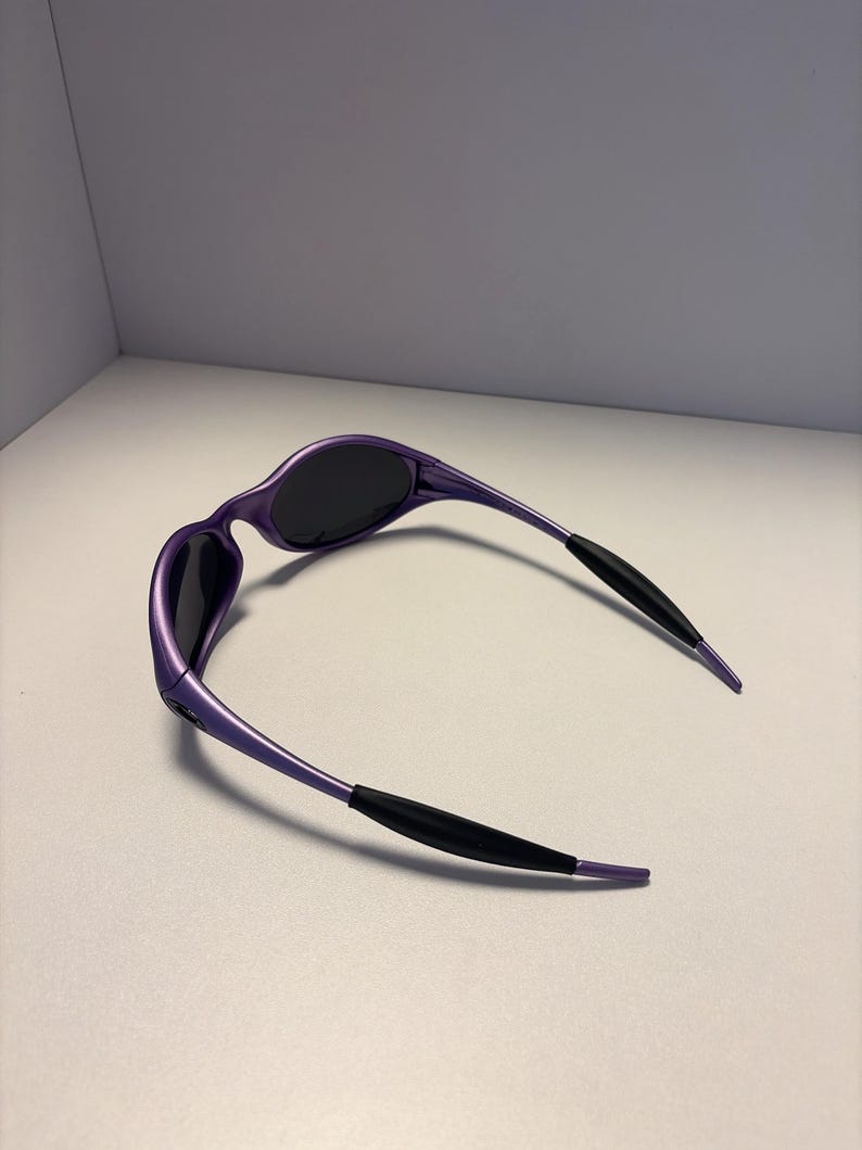 K&ouml;nnte beinhalten: Violette Sonnenbrille mit dunklen Gl&auml;sern und schwarzen B&uuml;geln. Die Sonnenbrille hat ein schlankes, geschwungenes Design. Die Fassungen sind in einem leuchtenden Violett gehalten, vor einem neutralen Hintergrund.