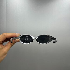 Pode incluir: &Oacute;culos de sol com arma&ccedil;&atilde;o prateada e lentes escuras. As lentes ovais est&atilde;o em uma arma&ccedil;&atilde;o elegante e moderna. Os &oacute;culos de sol s&atilde;o exibidos em um fundo neutro, mostrando seu design.