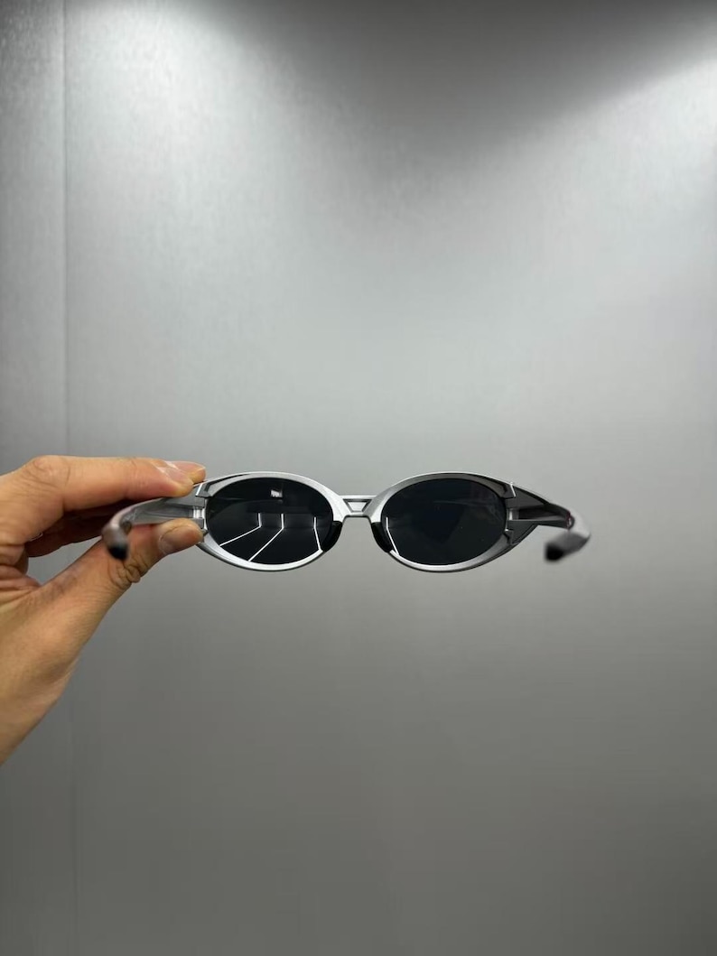 Pode incluir: &Oacute;culos de sol com arma&ccedil;&atilde;o prateada e lentes escuras. As lentes ovais est&atilde;o em uma arma&ccedil;&atilde;o elegante e moderna. Os &oacute;culos de sol s&atilde;o exibidos em um fundo neutro, mostrando seu design.