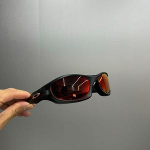 May include: Black sunglasses with red lenses. The sunglasses have a sleek, curved design and a matte finish. The lenses reflect the surrounding environment. The frame features a small logo on the temple.