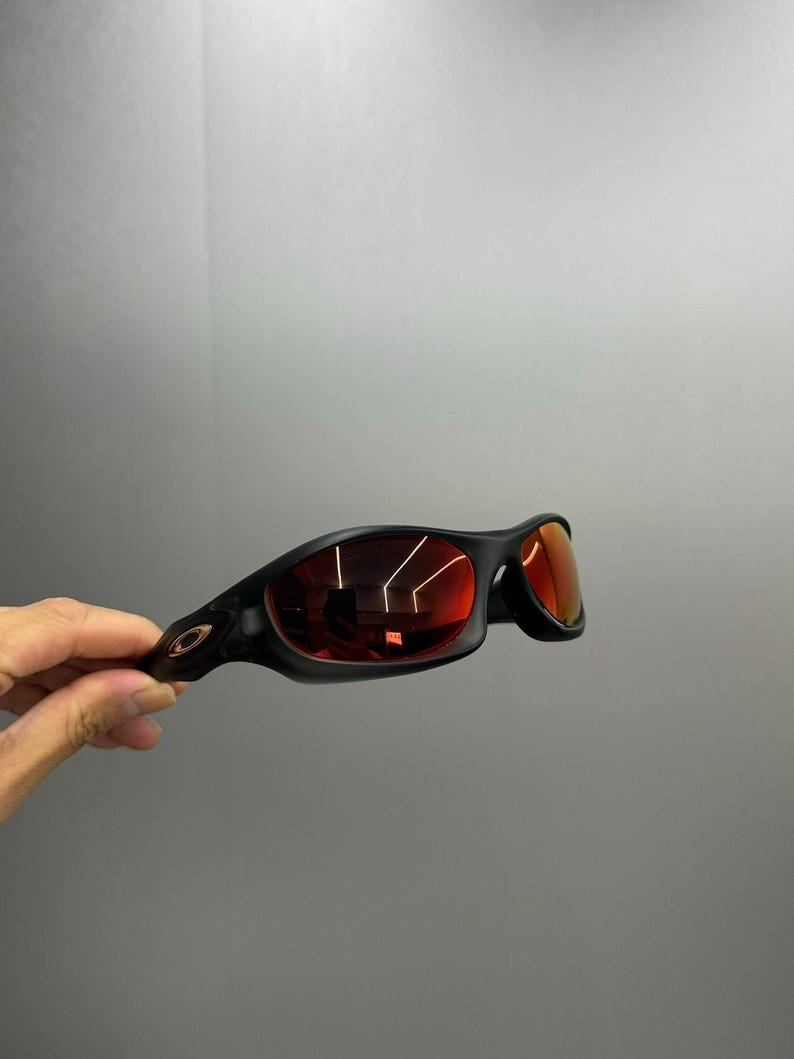 May include: Black sunglasses with red lenses. The sunglasses have a sleek, curved design and a matte finish. The lenses reflect the surrounding environment. The frame features a small logo on the temple.