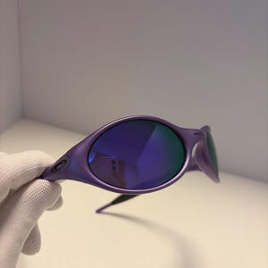 May include: Purple sunglasses with blue mirrored lenses. The sunglasses have a sleek, modern design with a curved frame and a small logo on the temple. The lenses reflect the surrounding environment. The sunglasses are held by a gloved hand.