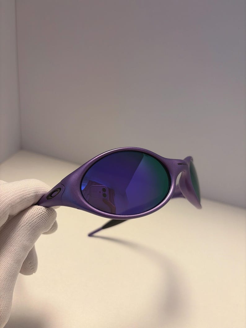 May include: Purple sunglasses with blue mirrored lenses. The sunglasses have a sleek, modern design with a curved frame and a small logo on the temple. The lenses reflect the surrounding environment. The sunglasses are held by a gloved hand.