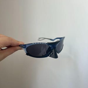 May include: Blue sunglasses with dark lenses. The sunglasses have a sporty design with a nose guard and a textured frame. The arms of the sunglasses are a lighter shade of blue.