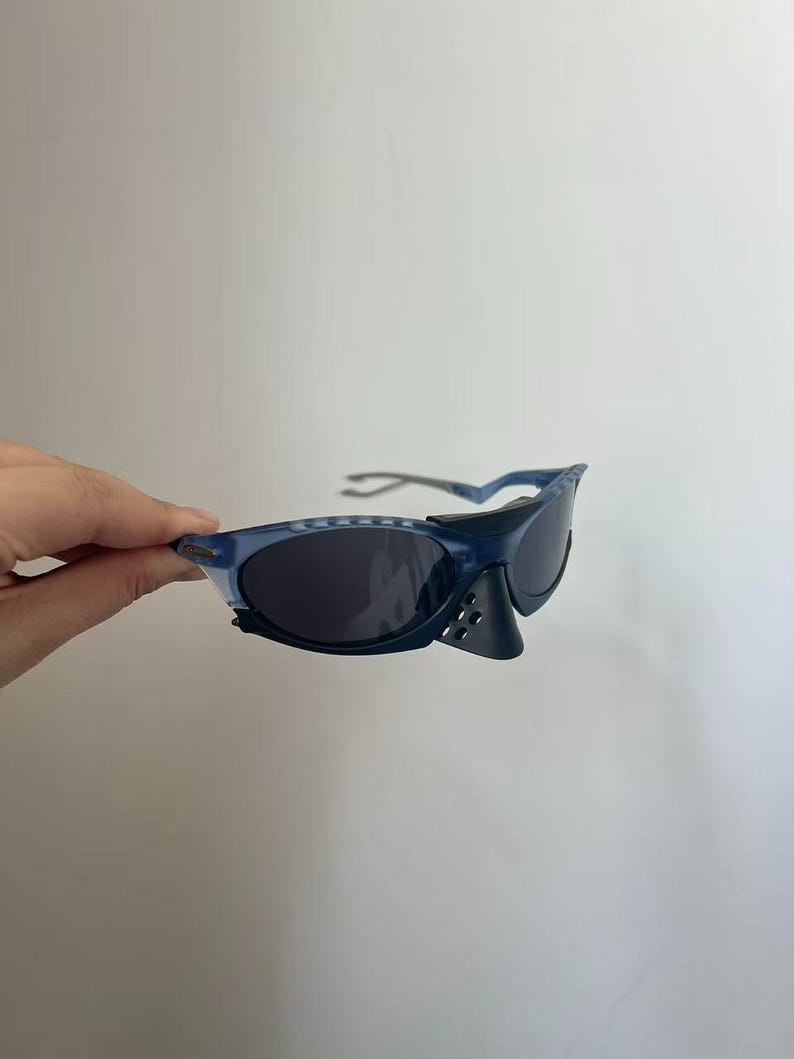 May include: Blue sunglasses with dark lenses. The sunglasses have a sporty design with a nose guard and a textured frame. The arms of the sunglasses are a lighter shade of blue.