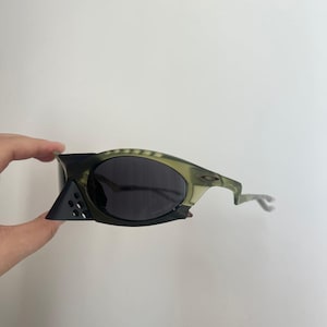 May include: A pair of olive green and black sunglasses with dark lenses. The sunglasses have a unique design with a geometric shape on the side and a curved arm. The lenses are dark, and the frame is a mix of green and black.