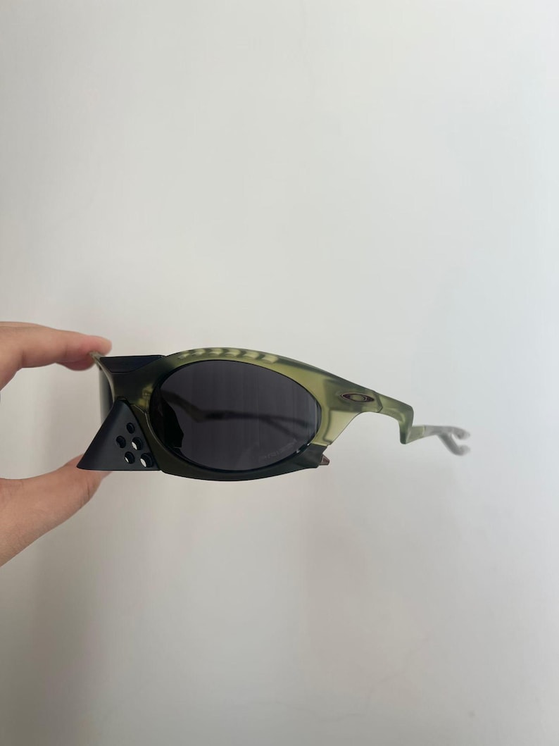 May include: A pair of olive green and black sunglasses with dark lenses. The sunglasses have a unique design with a geometric shape on the side and a curved arm. The lenses are dark, and the frame is a mix of green and black.