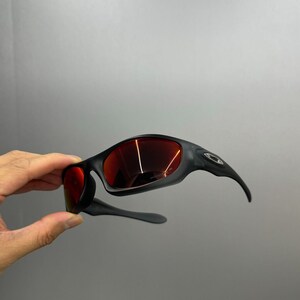 May include: Black sunglasses with red lenses. The sunglasses have a sleek, curved design with a matte finish. The lenses reflect light, and the arms curve down towards the ears. The Oakley logo is visible on the side.